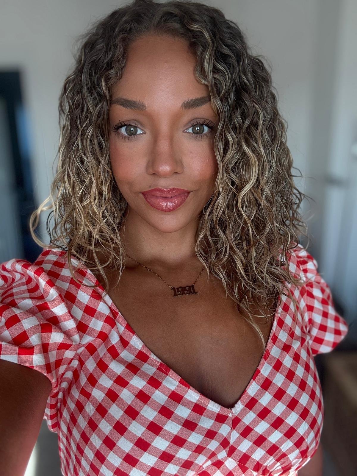 Close-up portrait of a smiling woman with curly hair, wearing a red and white checkered top and a necklace that says "Real."