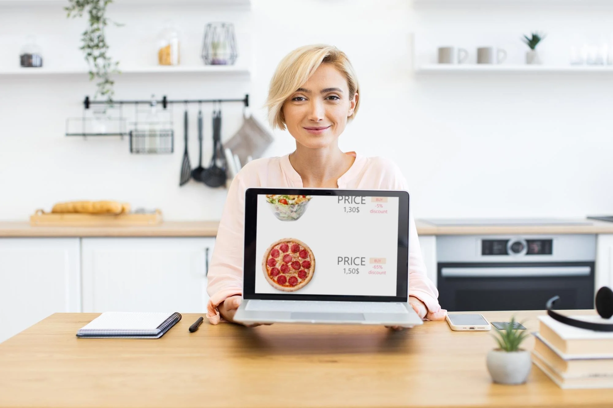 Woman in a light pink shirt smiling and holding a laptop showing images of a salad and a pizza with prices and discounts, sitting at a wooden table in a bright white kitchen.