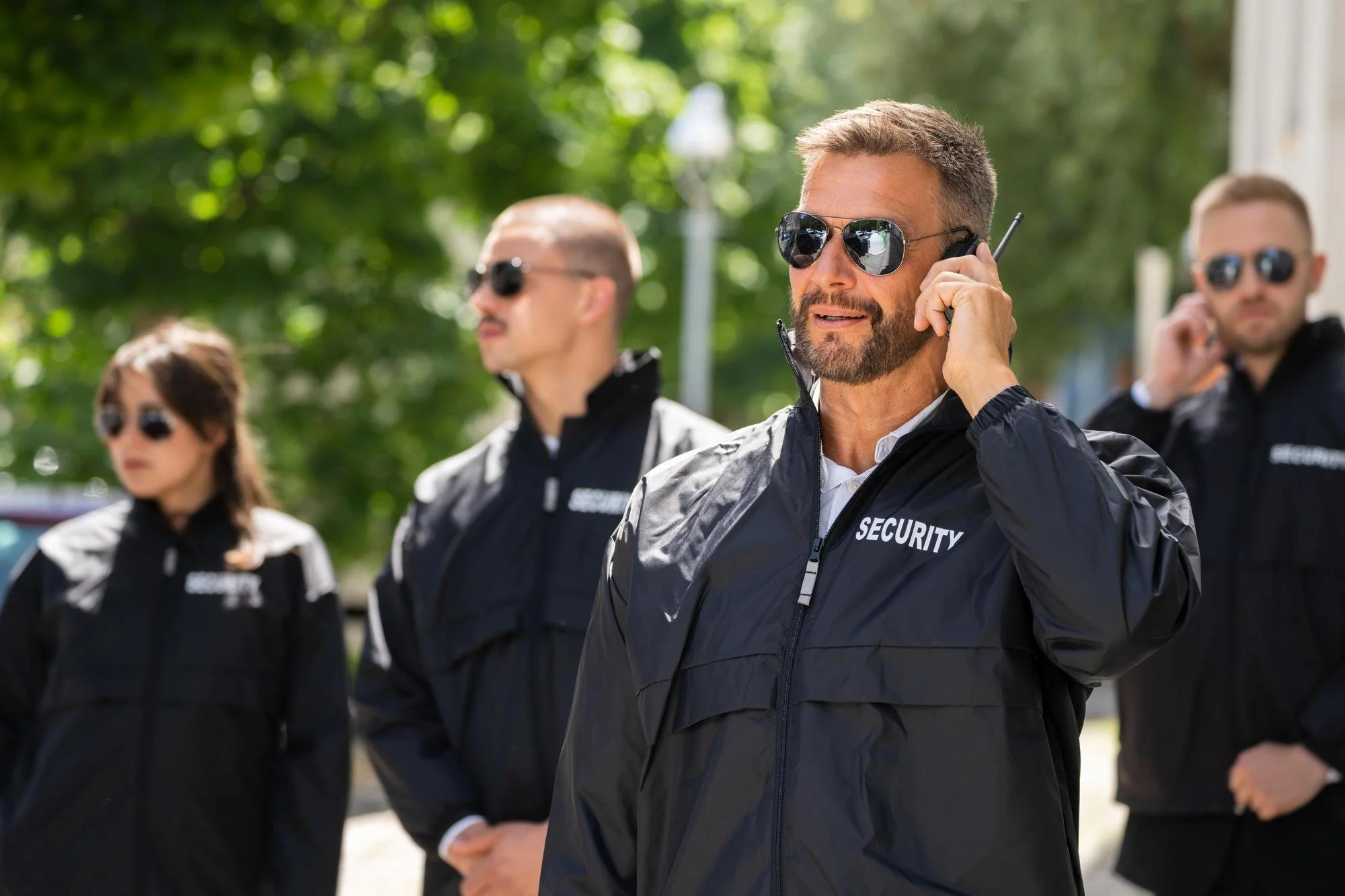 A group of security personnel wearing black jackets with 'SECURITY' written on them, standing outdoors on a sunny day. One man in the foreground is speaking on a walkie-talkie and wearing sunglasses and a beard. Three other security staff members are standing behind him, also wearing sunglasses.