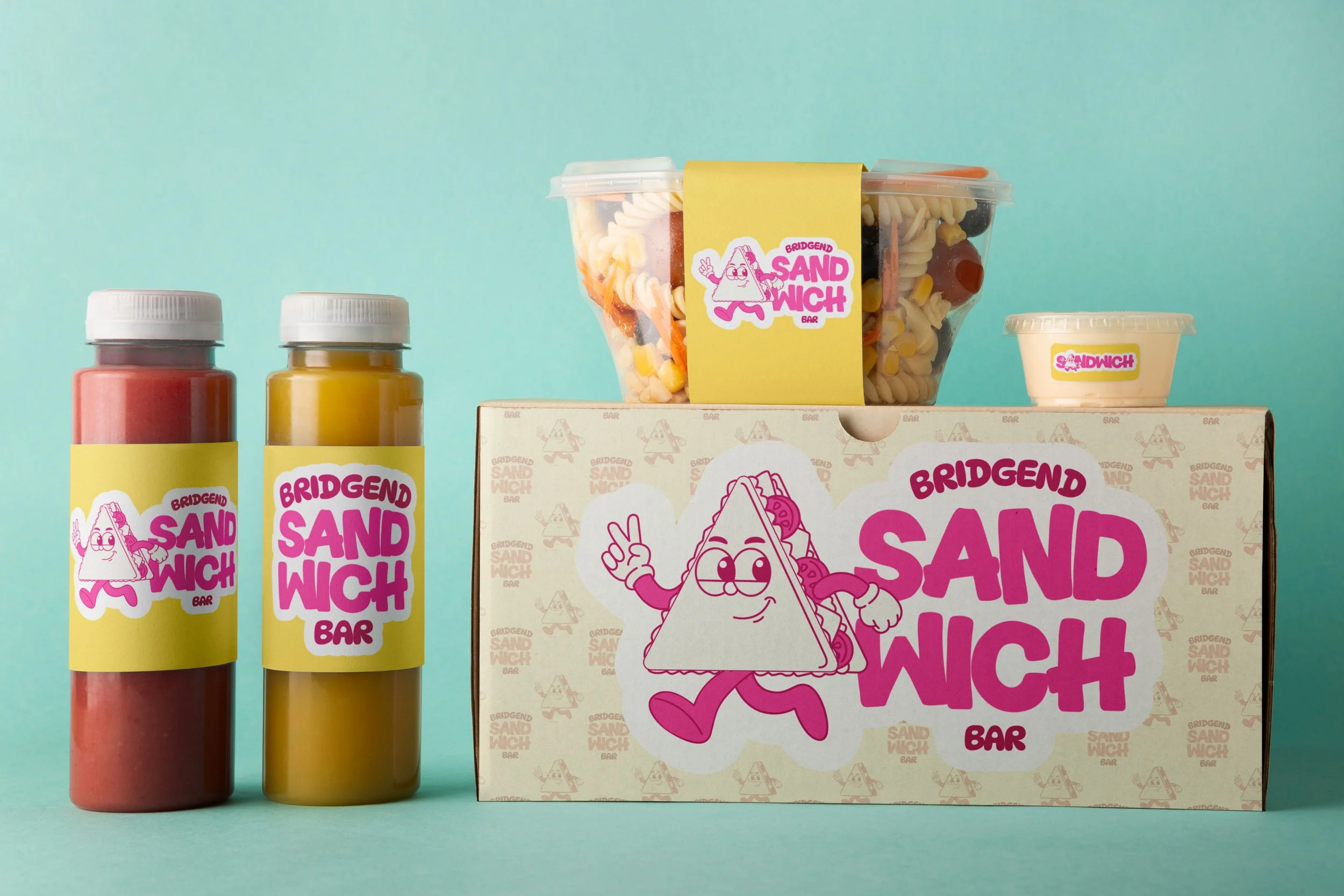 A box labeled 'Bridgend Sand Wich Bar' with cartoon illustrations of a smiling sandwich character making a peace sign, with two bottles of condiment in front and a cup of sandwich toppings or spread on the right, all set against a pastel background.