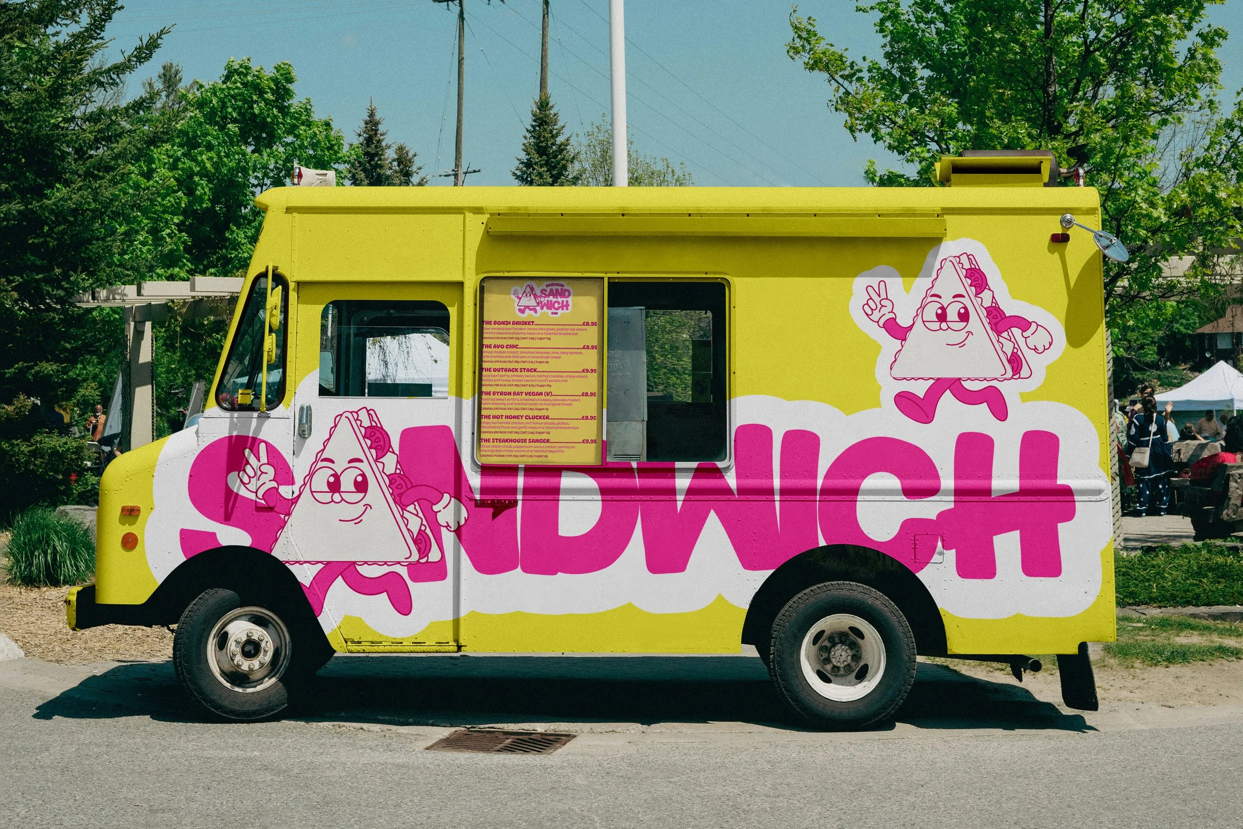 A yellow food truck featuring a cartoon character of a smiling sandwich with glasses, making a peace sign, and a matching sandwich mascot on the side with pink accents, parked outdoors with trees and cloudy sky in the background.