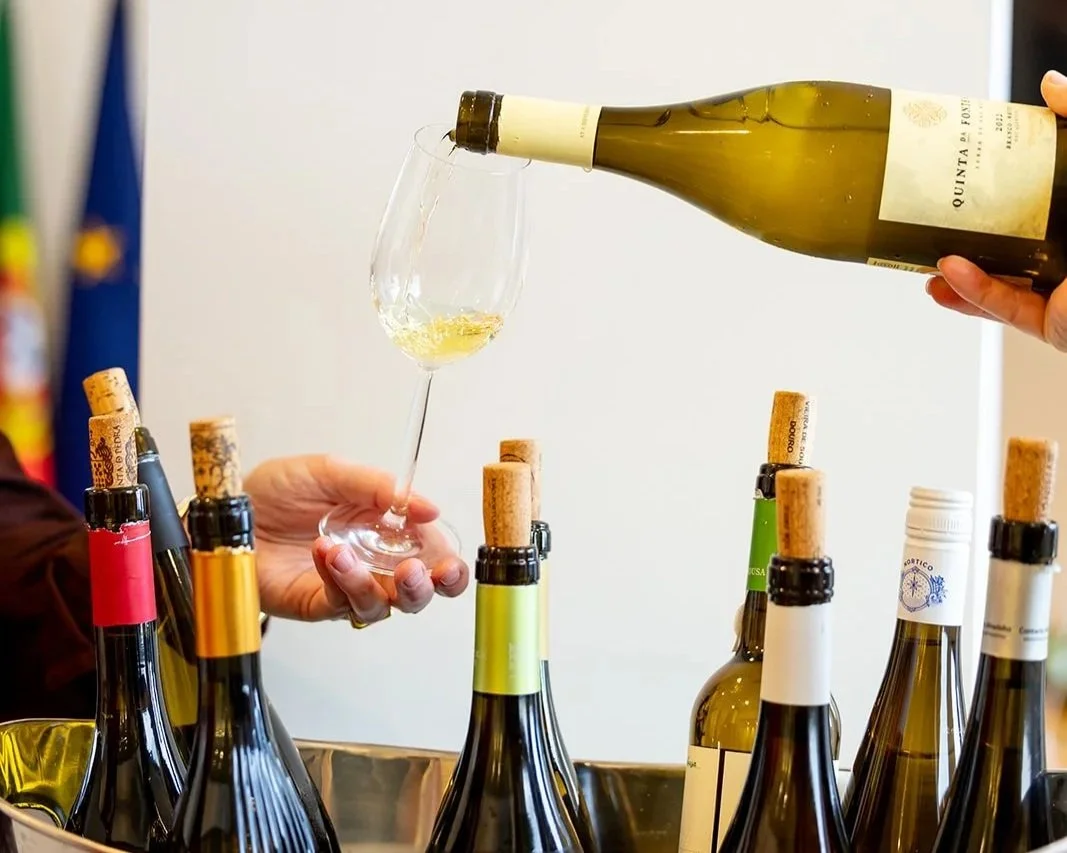 Person pouring white wine into a glass at a wine tasting event with several wine bottles on a table.