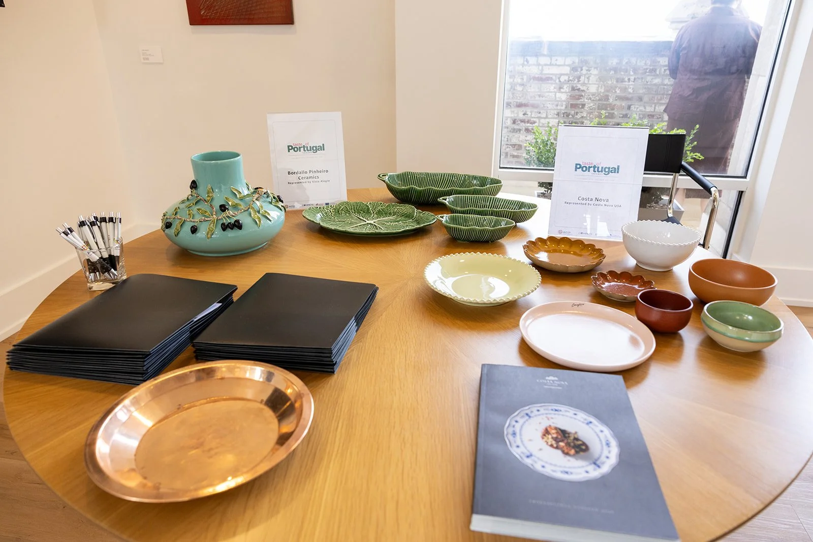 Display of colorful ceramic bowls and plates on a wooden table, with signs indicating they are from Portugal, including labels for Bordallo Pinheiro Ceramics and Costa Nova, and black notebooks, pens, and a copper tray.