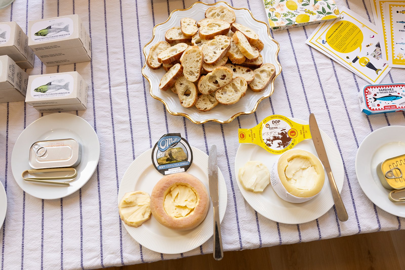 A table with sliced bread on a decorative plate, cheese, butter, canned fish, and small cartons of milk or juice. There are also plates with cheese and utensils, and some informational cards.