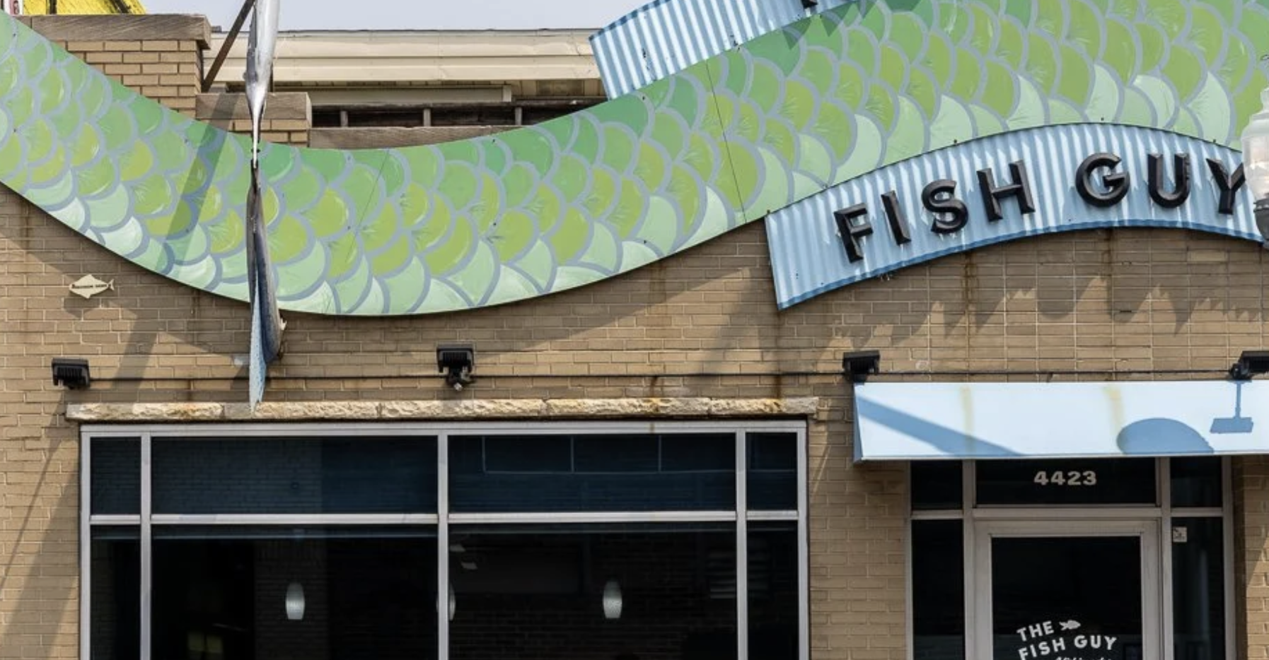 Exterior of a building with a large decorative sign featuring a green fish scale pattern and the text 'FISH GUY', along with a smaller sign for 'THE FISH GUY' business at the entrance. The building has a brick facade, large windows, and the address number 4423.