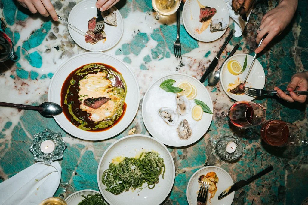 A dinner table at a restaurant with dishes including oysters on a plate with lemon slices, green spaghetti, a bowl of creamy sauce, and a plate with a piece of meat. There are also glasses of red and white wine, candles, and utensils.