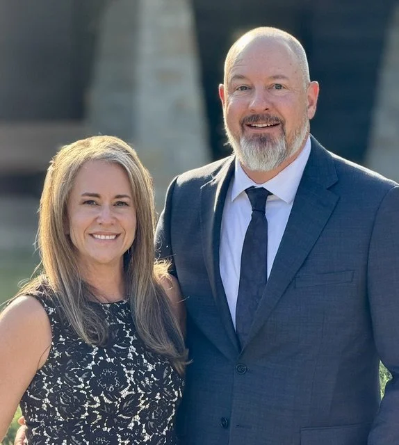 A smiling woman with long blonde hair wearing a black and white lace dress standing next to a smiling man with a beard in a blue suit and tie, outdoors with blurred buildings in the background.
