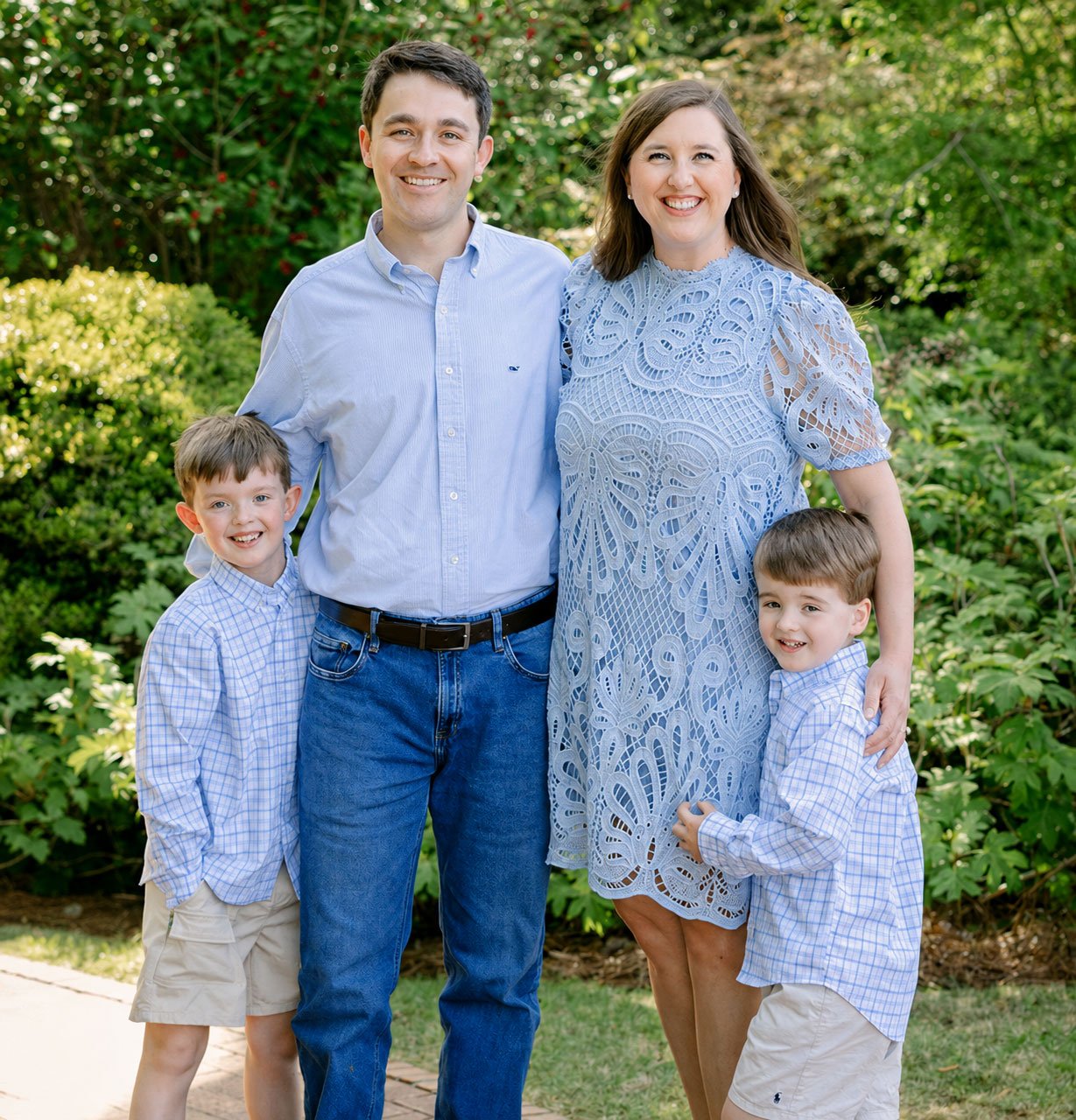 A happy family of four standing outdoors, smiling at the camera. The father and mother are in the center, with two young boys on either side. The background features greenery and sunlight filtering through trees.