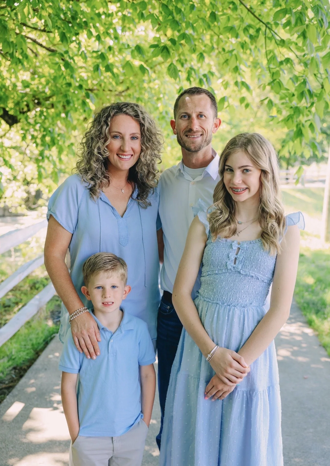 A family of five posing outdoors on a sunny day, surrounded by green trees and a white fence, all dressed in shades of blue.