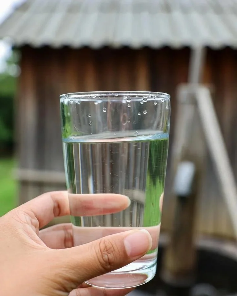 A hand holding a glass of water outdoors with a wooden structure and greenery in the background.