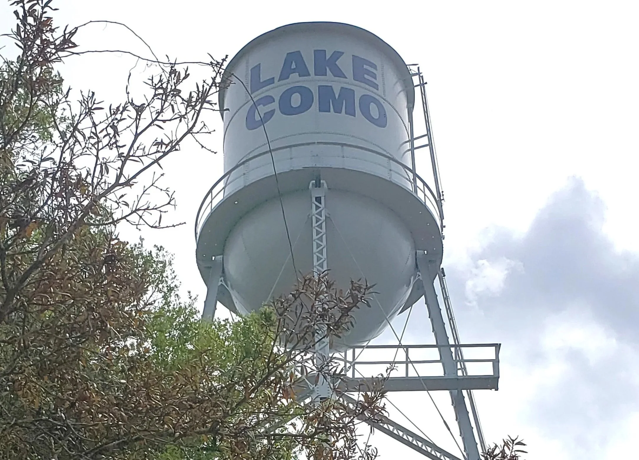 A tall white water tower with the words 'LAKE COMO' written on it in large blue letters, situated above a tree with some cloudy sky in the background.