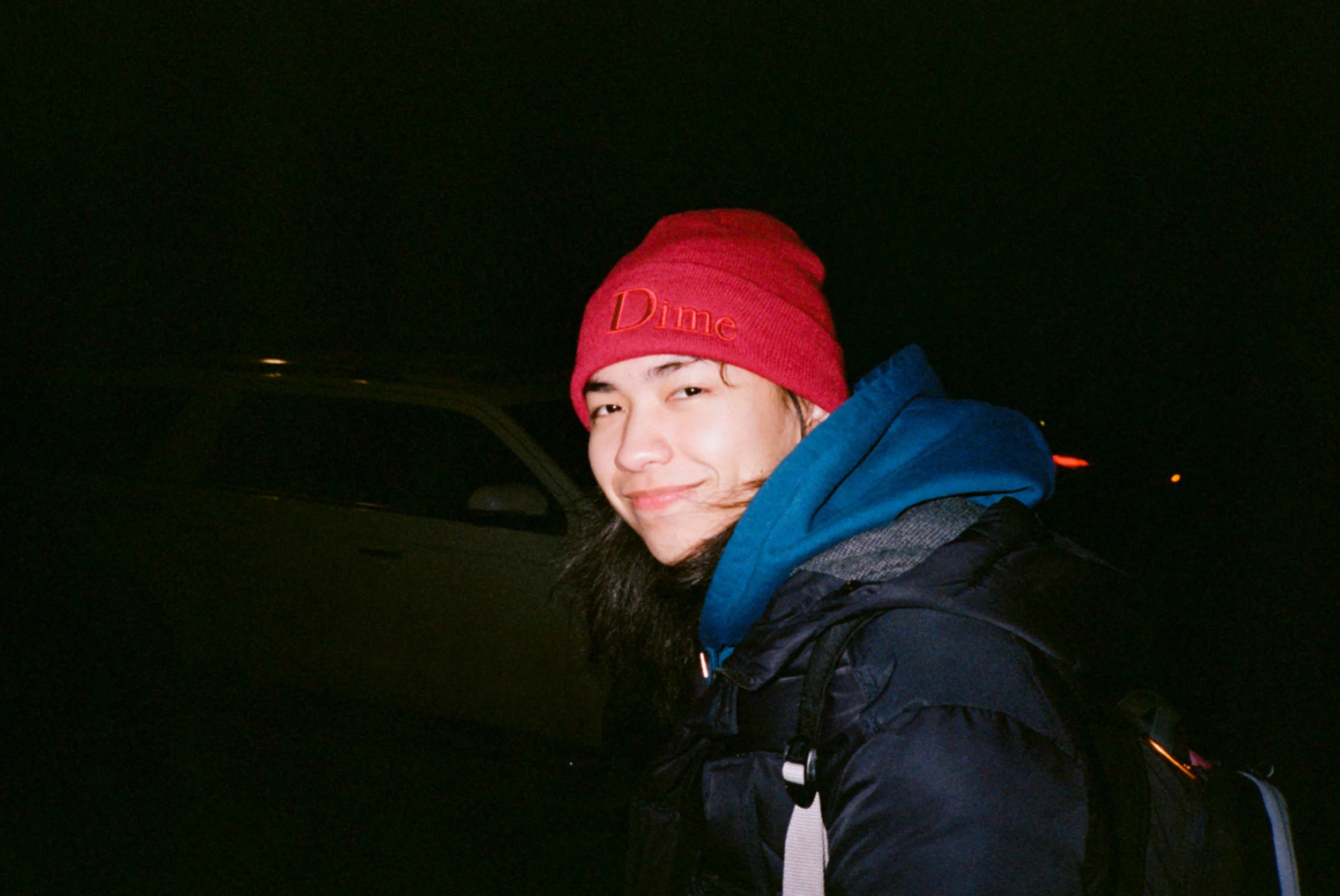 A person smiling at night, wearing a red beanie with the word 'Dime' embroidered on it and a blue jacket.