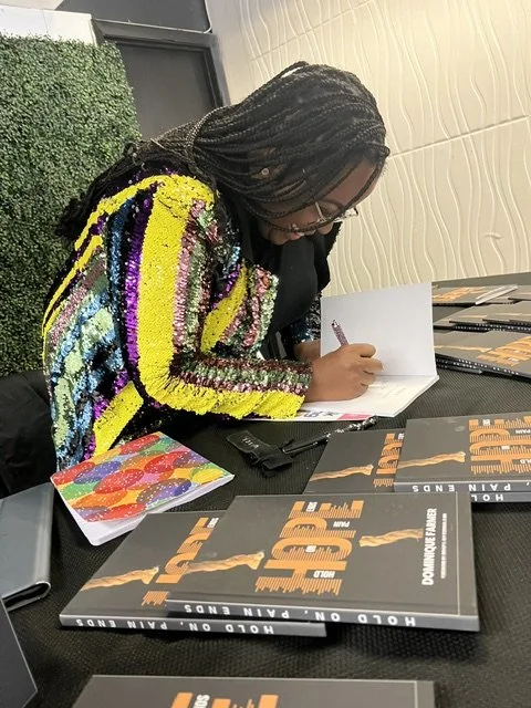 Person with dreadlocks wearing a colorful, sequined jacket signing a book at a table.