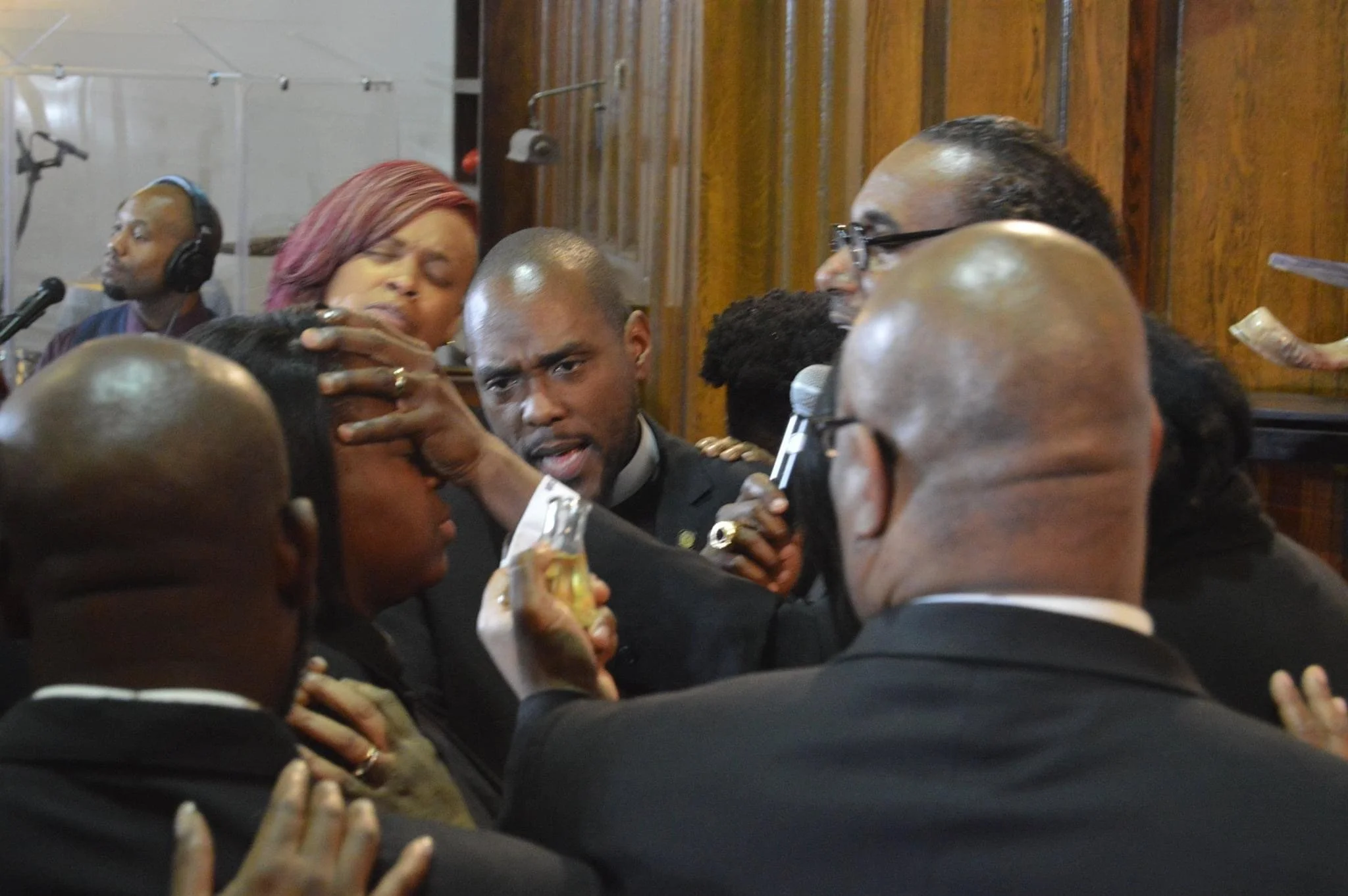 A group of people dressed in formal attire receiving blessings from a man holding a microphone during a ceremony.