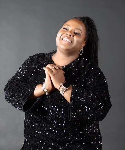 A woman smiling and looking upward, wearing a black sequin jacket and jewelry, against a gray background.