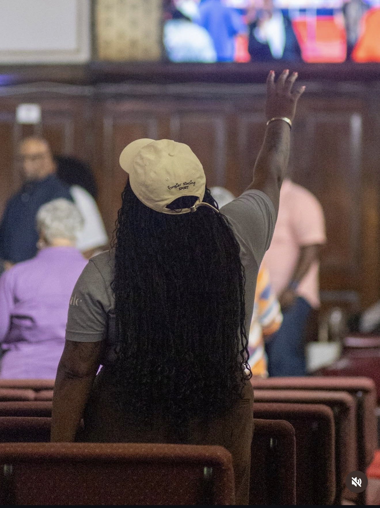 A woman with long, curly black hair wearing a beige cap with black details and a gray shirt, standing with her back to the camera, reaching out with her right arm towards a television screen in a room filled with people.