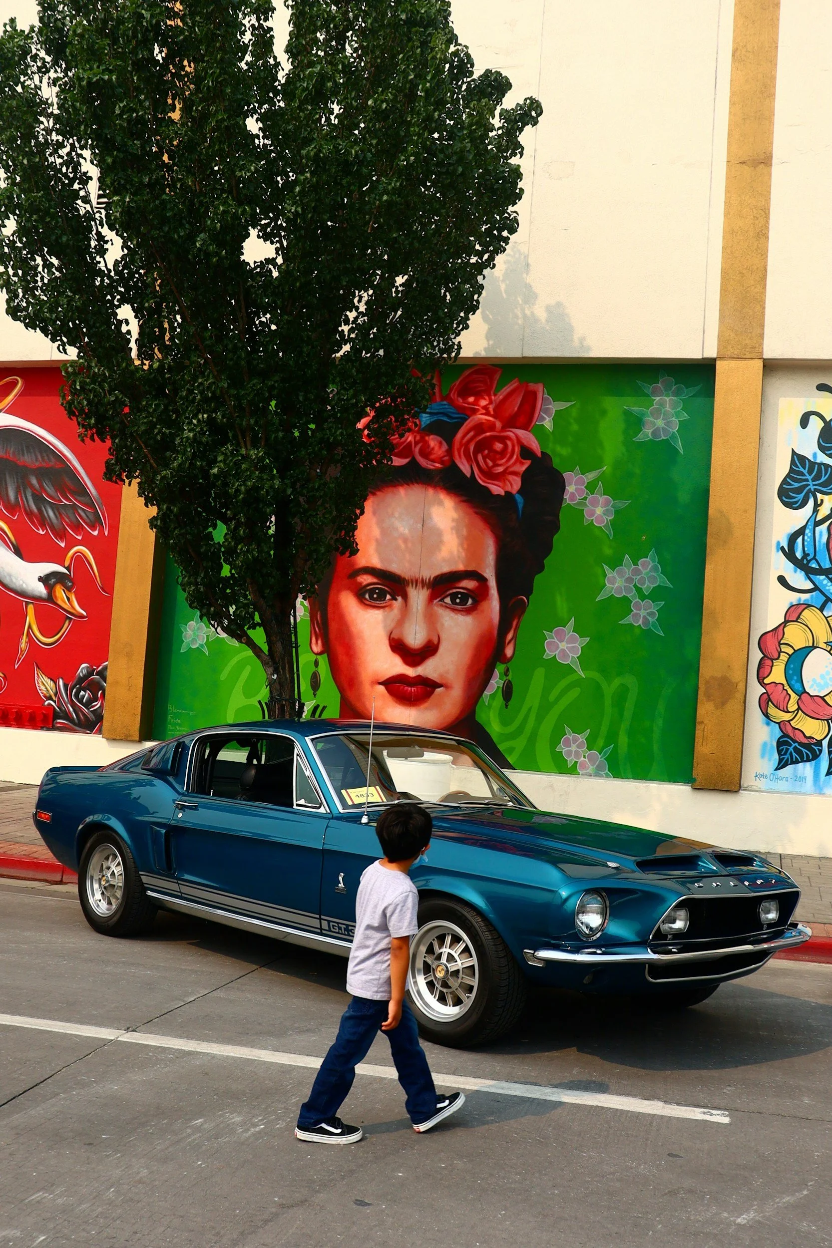 A vintage blue Ford Mustang car parked on the street with a boy walking past in the foreground. Behind the car are murals on the wall, including a woman with flowers in her hair and artwork of other colorful figures. A tree partly covers the murals.