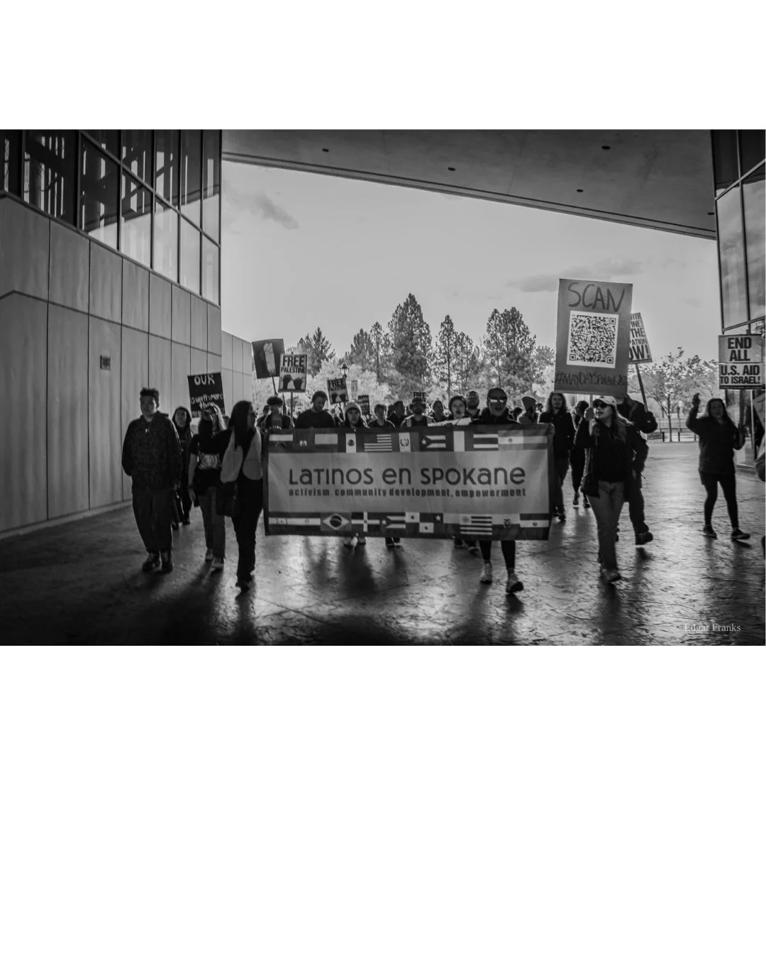 Black and white photo of a diverse group of protesters marching outdoors, holding signs and a banner that reads 'Latinos en Spokane' with flags from various countries, advocating for activism, community development, and empowerment.