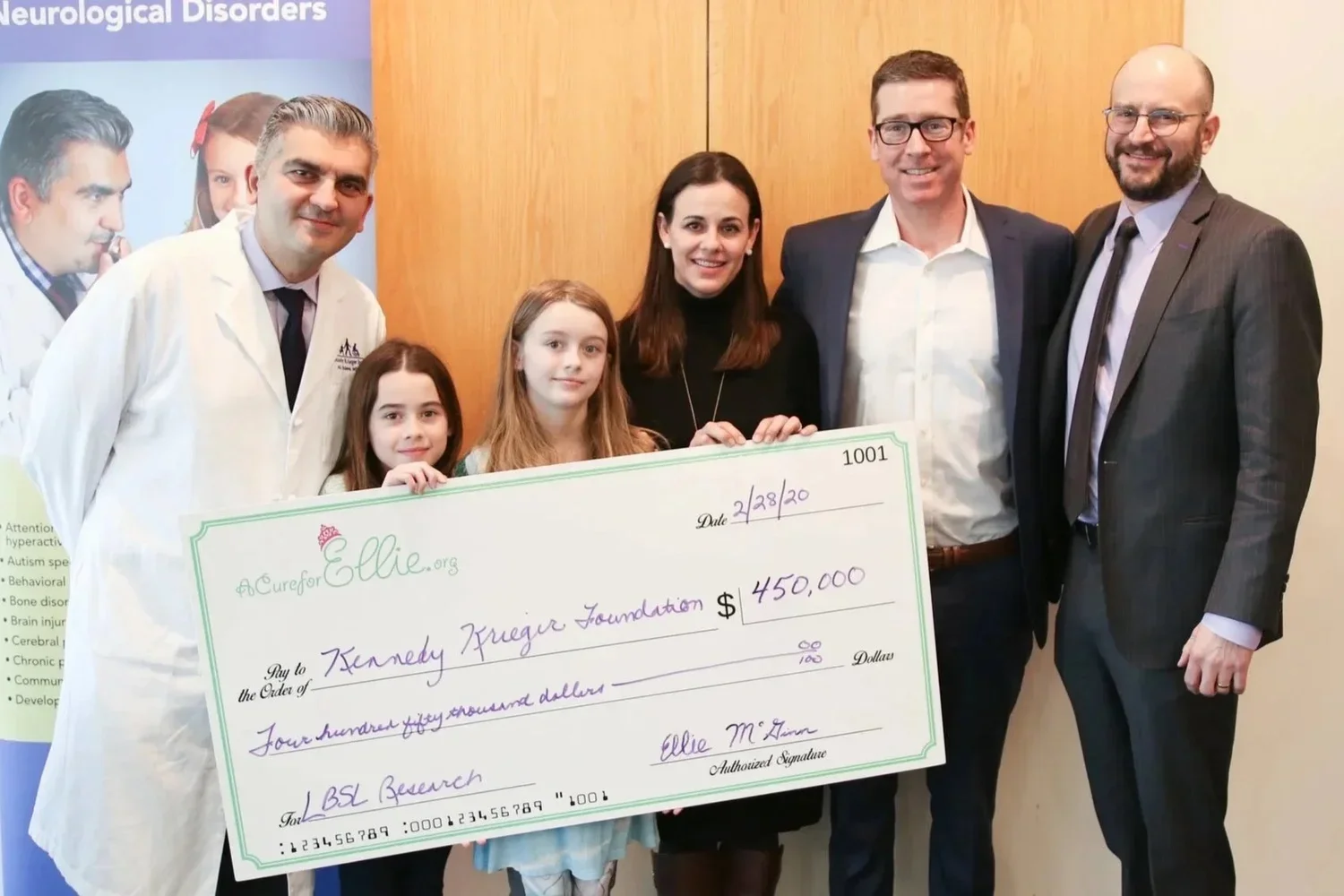 Group of people holding a large donation check for $450,000 made out to the Kennedy Krieger Foundation, with children and medical professionals present.