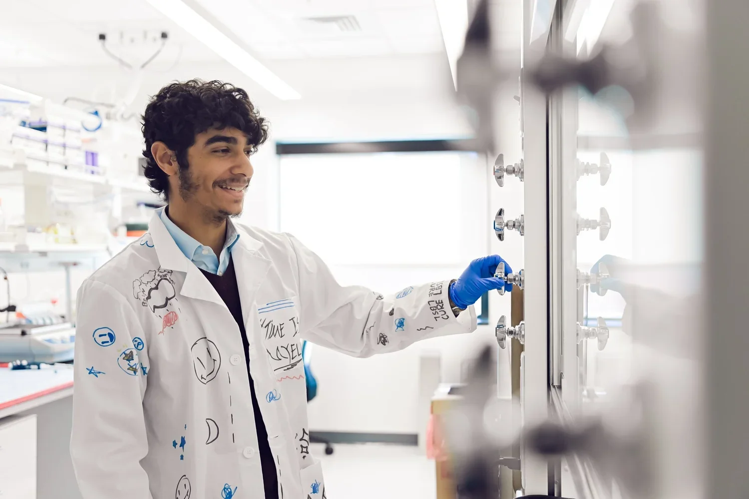 A young male scientist wearing a white lab coat with doodles and words, blue gloves, and a collared shirt, working in a laboratory and adjusting a knob on a control panel.