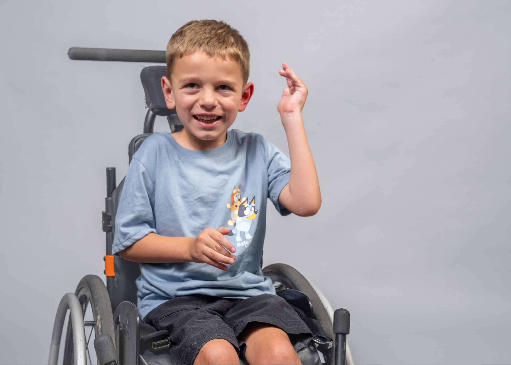A young boy with short light brown hair, smiling and raising his right hand, sitting in a wheelchair against a plain light gray background. He is wearing a light blue t-shirt with cartoon characters on it and dark shorts.