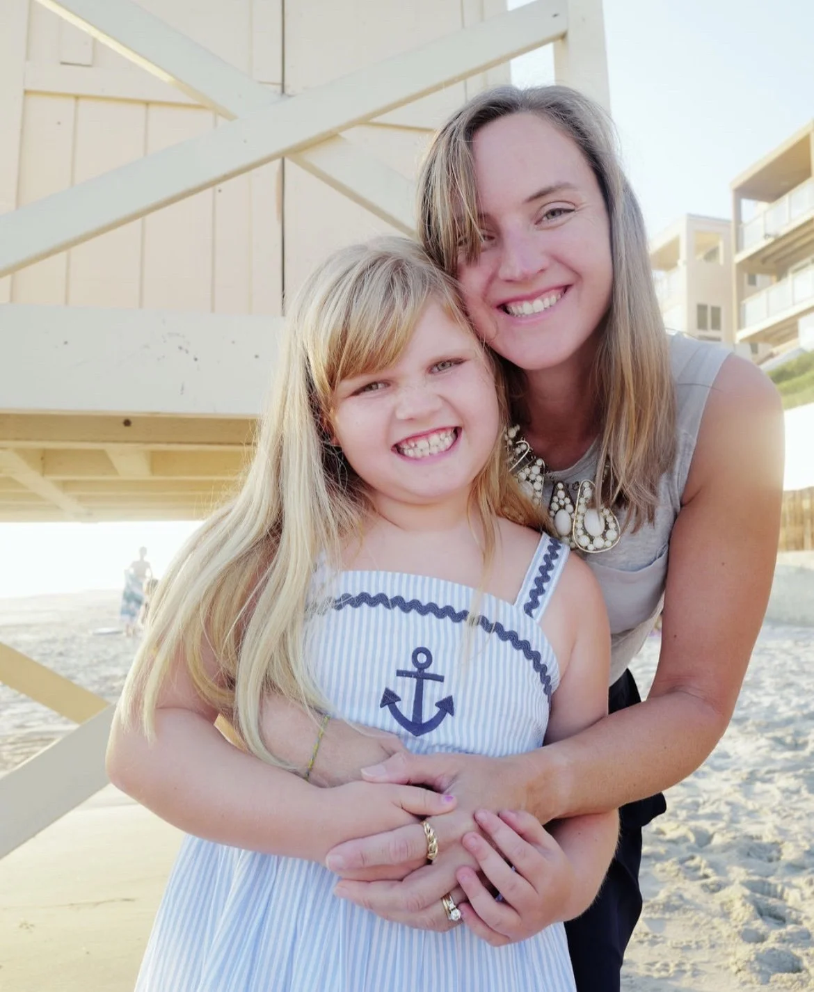 A woman and a young girl smiling and hugging at the beach, with a lifeguard stand and buildings in the background.