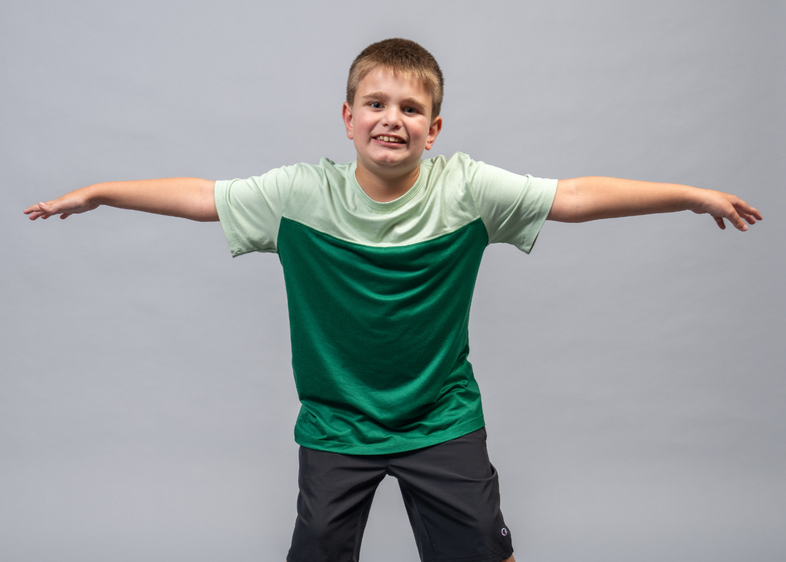A young boy in a green shirt and black pants stands with his arms stretched out