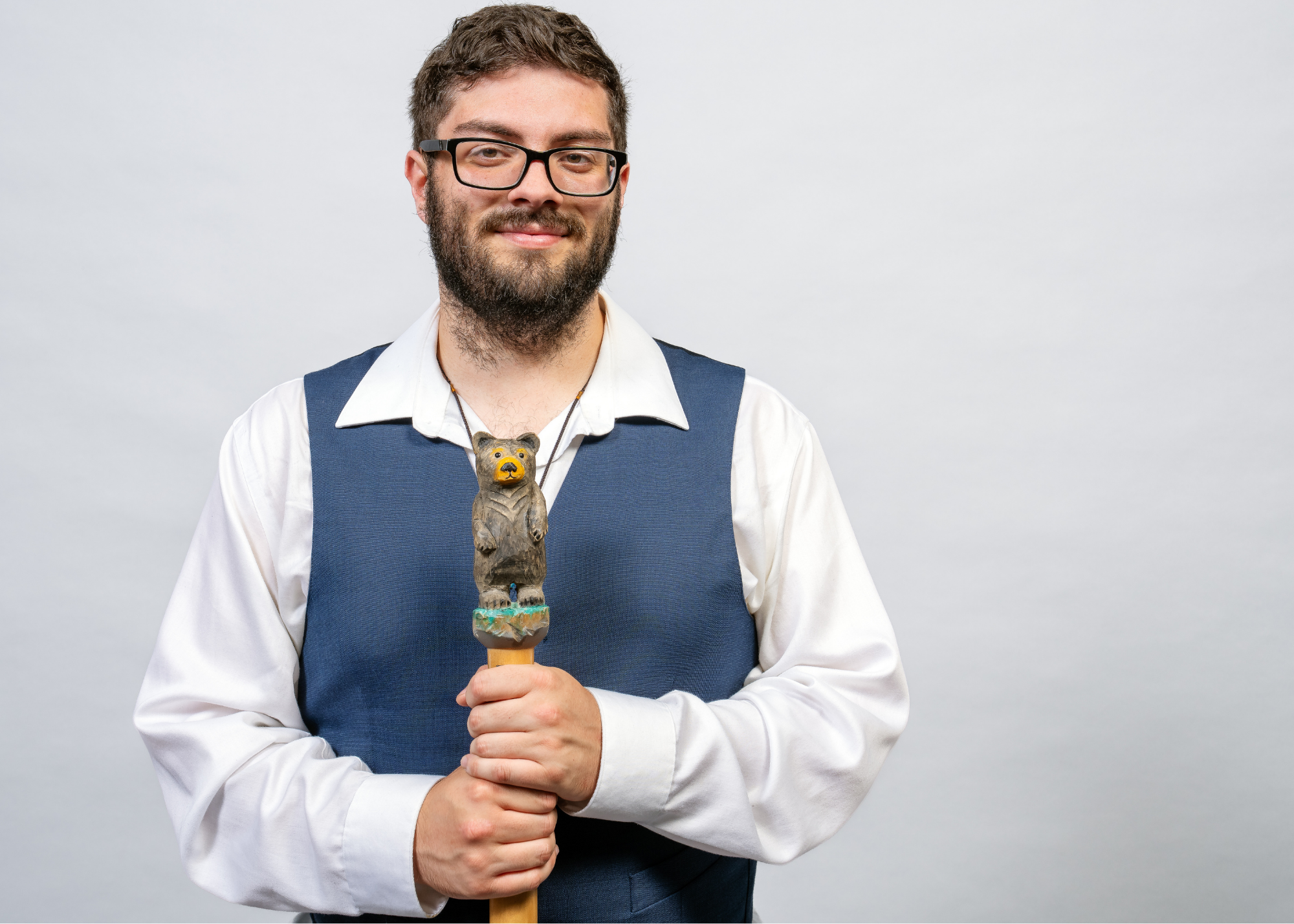 A man in a white shirt and a blue vest holding a carved walking stick with a bear on top