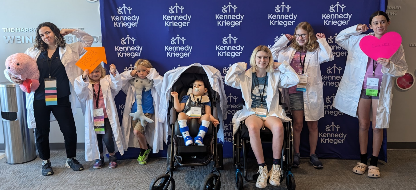 Group of children and a woman posing in front of a blue Kennedy Krieger backdrop, wearing white lab coats with some holding toys and signs, and two children in wheelchairs at the center.