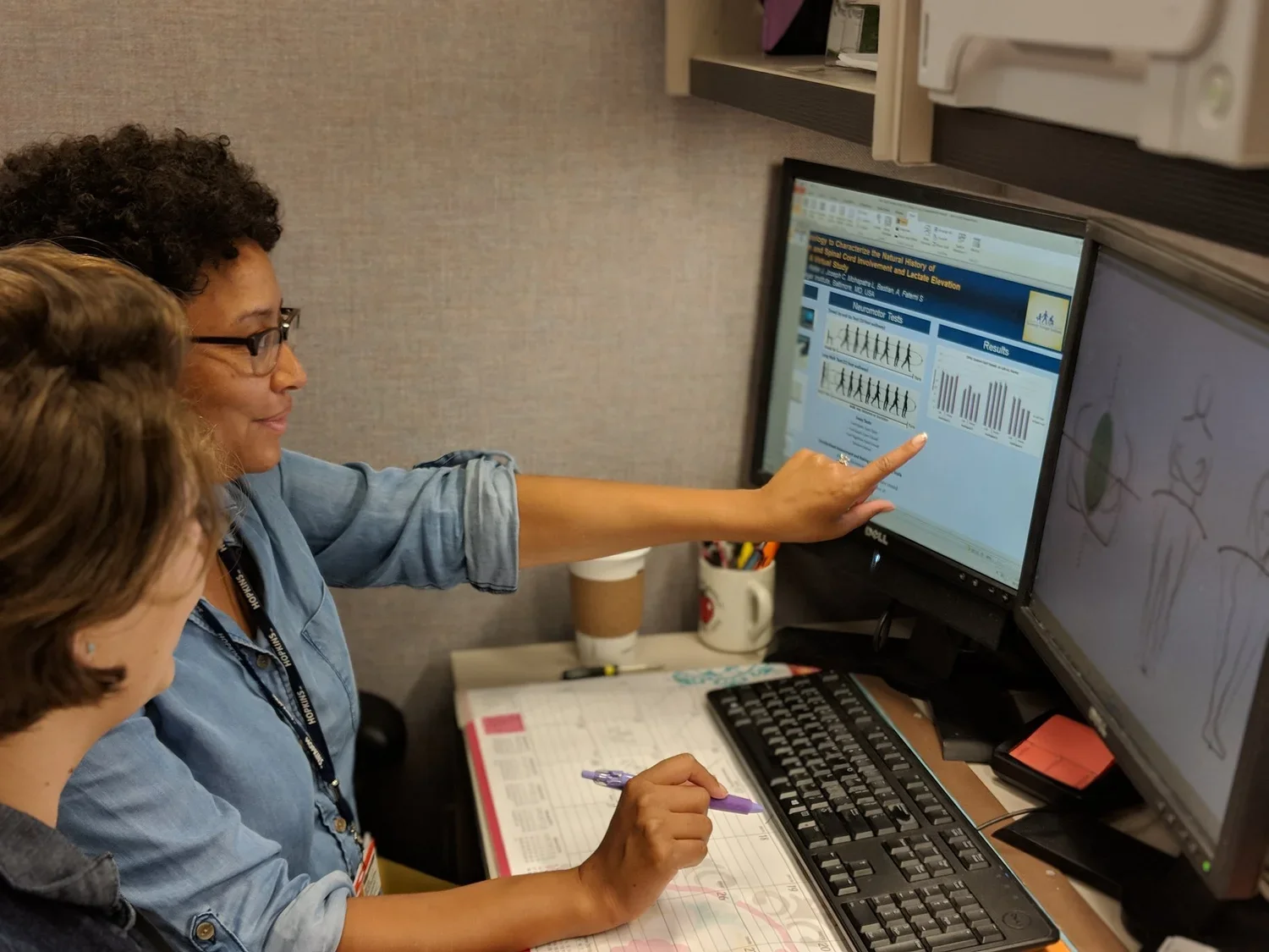 Two women working at a desk with dual computer monitors, one showing a presentation with charts and graphs, the other displaying drawings of people.