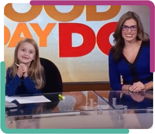 Two women sitting at a news desk with a large TV screen behind them displaying the words 'Good Day D.C.'