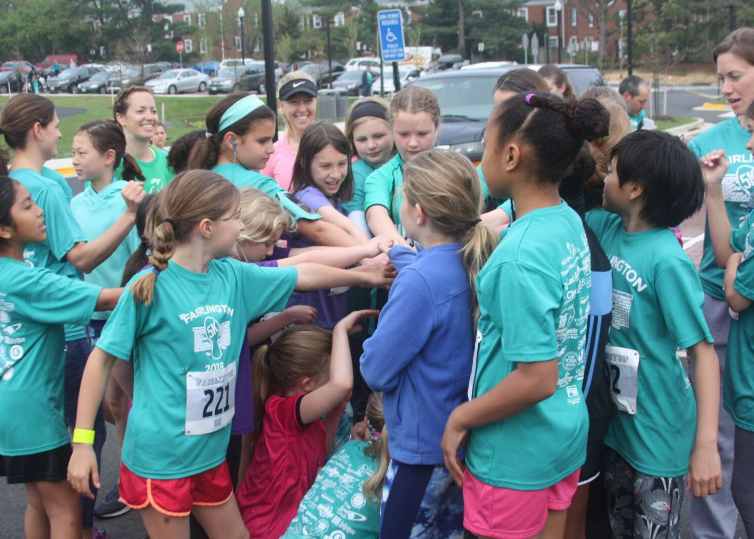 A group of people, mostly young girls and women, wearing teal shirts with event logos, huddled together in a circle outdoors, possibly indicating team spirit or unity at a race or event.