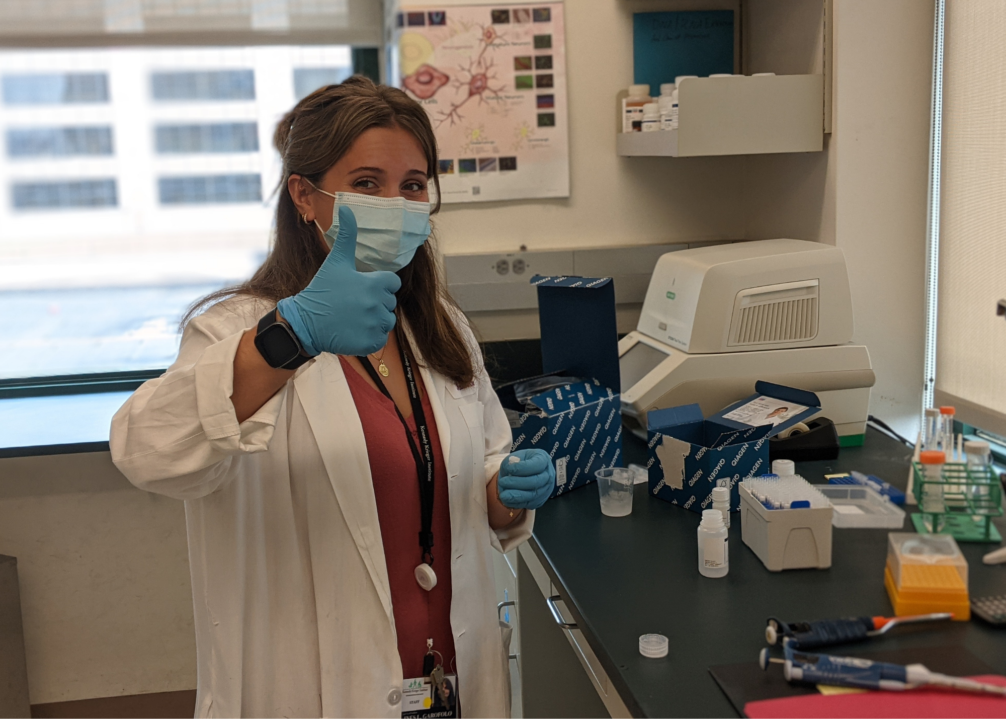 A female healthcare professional wearing a face mask and gloves, giving a thumbs-up in a laboratory or medical setting with scientific supplies and equipment on the counter.