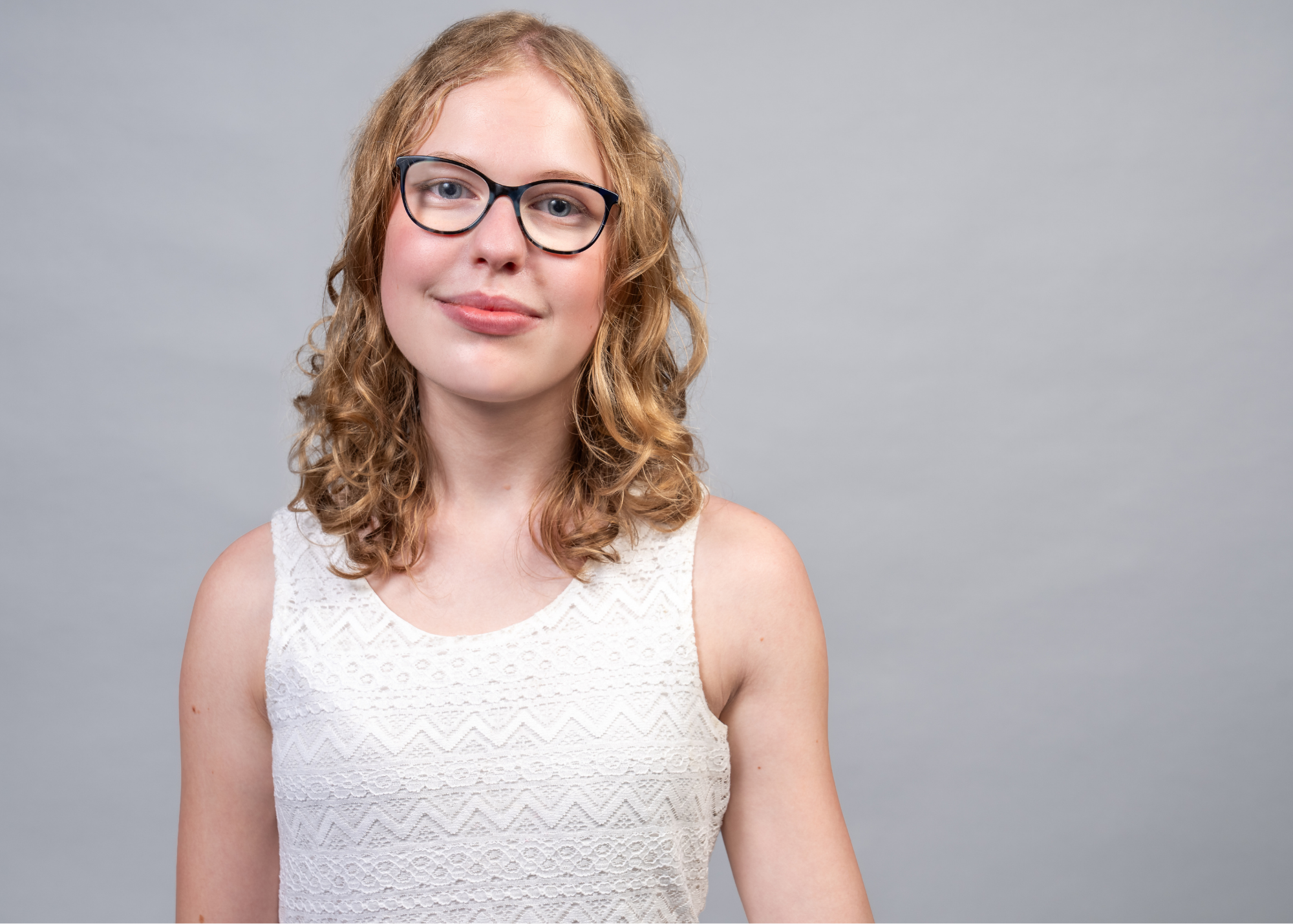 A young woman with curly blonde hair, wearing glasses and a white sleeveless lace top, standing against a plain gray background.