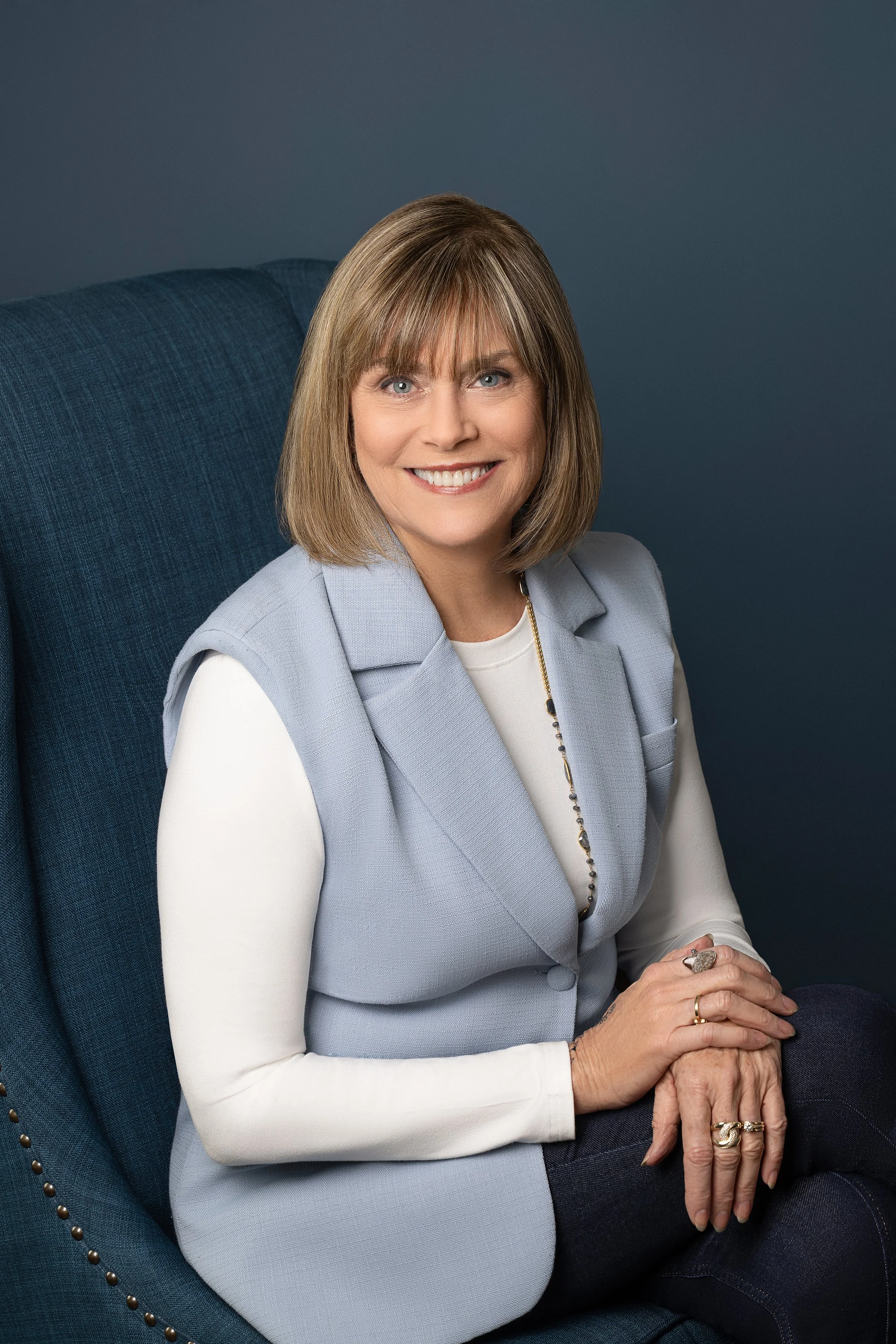 A middle-aged woman with blonde hair and blue eyes, smiling, sitting on a dark blue armchair, wearing a light gray vest over a white long-sleeve shirt, with rings on her fingers and a necklace, against a dark background.