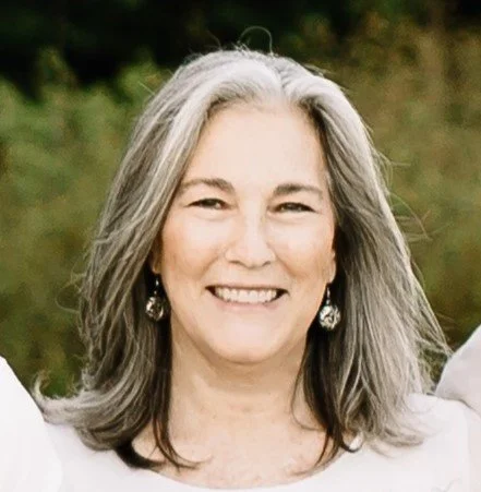 A woman with gray hair smiling outdoors, wearing earrings.