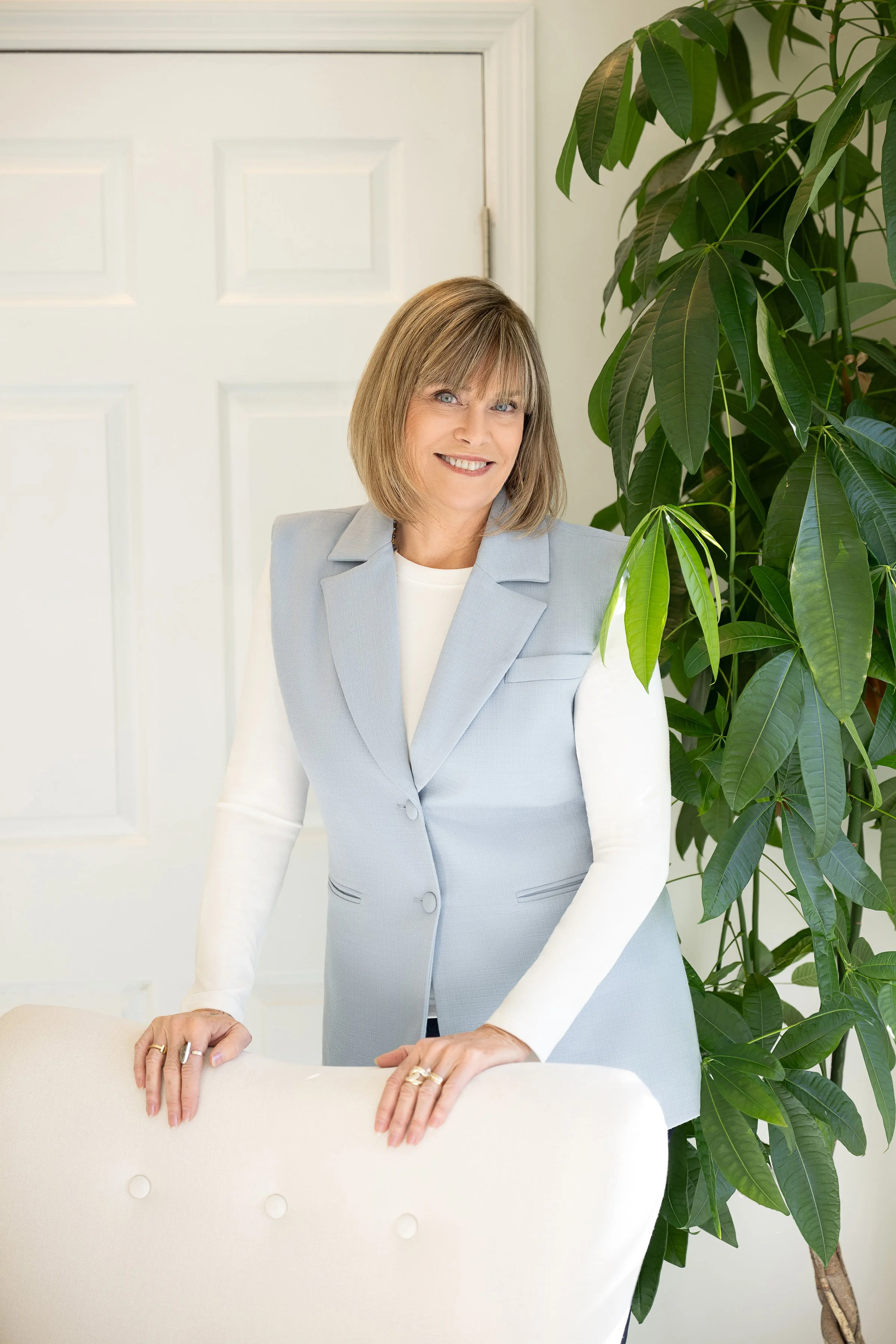 A woman with shoulder-length blonde hair and blue eyes, wearing a light gray blazer over a white shirt, smiling while leaning on the back of a white chair in a room with a white door and a large green plant.