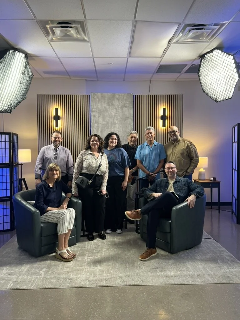 Group of eight people posing for a photo in a modern indoor setting with studio lights, decorative fixtures, and furniture. Some are standing, and two are seated in armchairs.