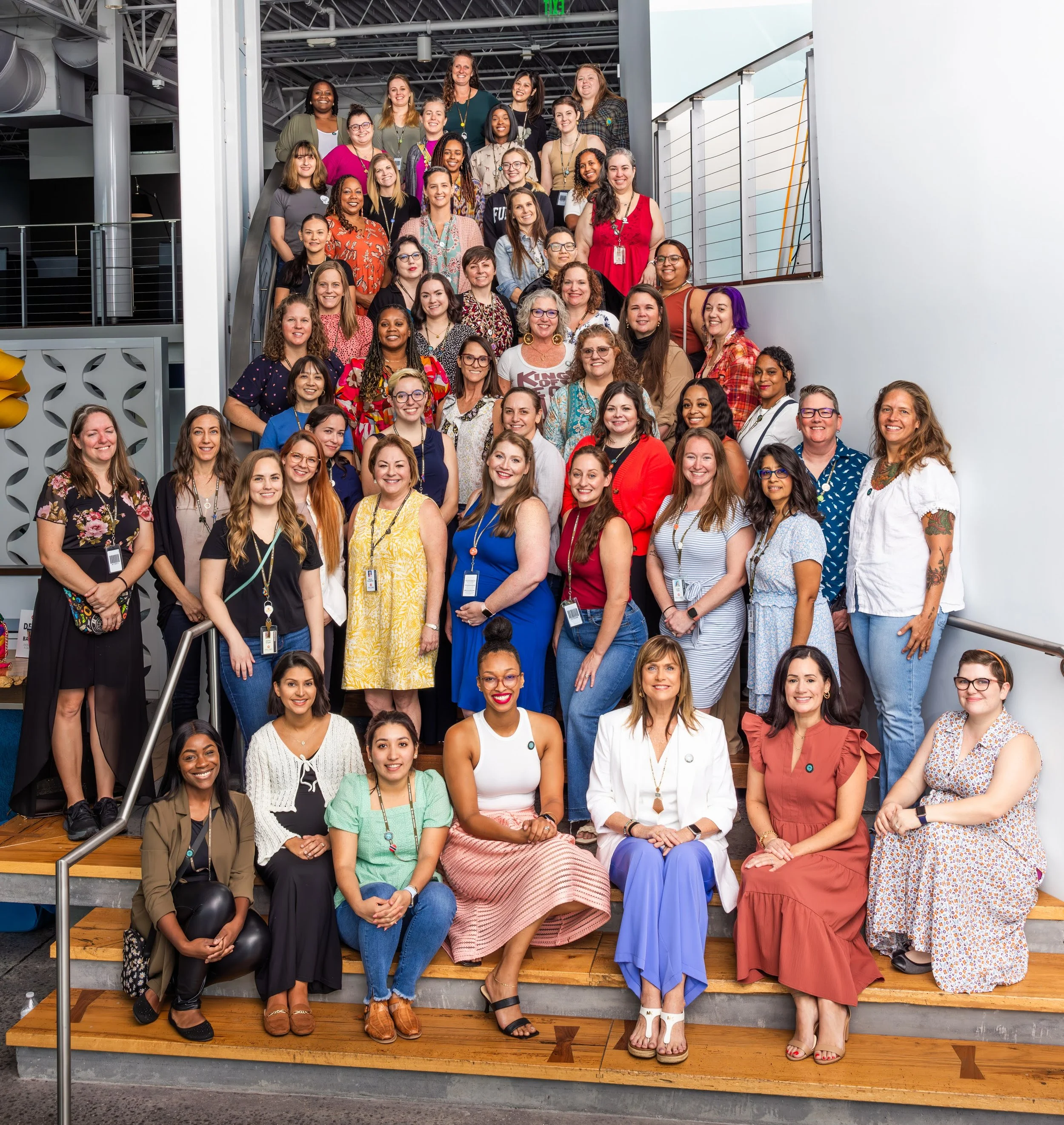 A group of diverse women smiling and posing on wooden stairs inside a modern building. The women are dressed in casual and colorful clothing, with some wearing glasses and name tags.