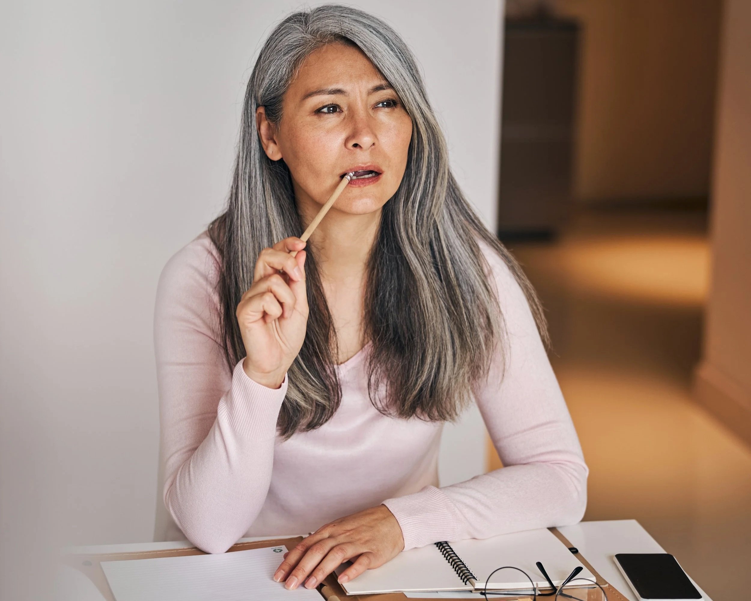 A middle-aged woman with long gray hair wearing a light pink long sleeve shirt, sitting at a white table, with a pen in her hand near her face, looking slightly to her right with a thoughtful expression.
