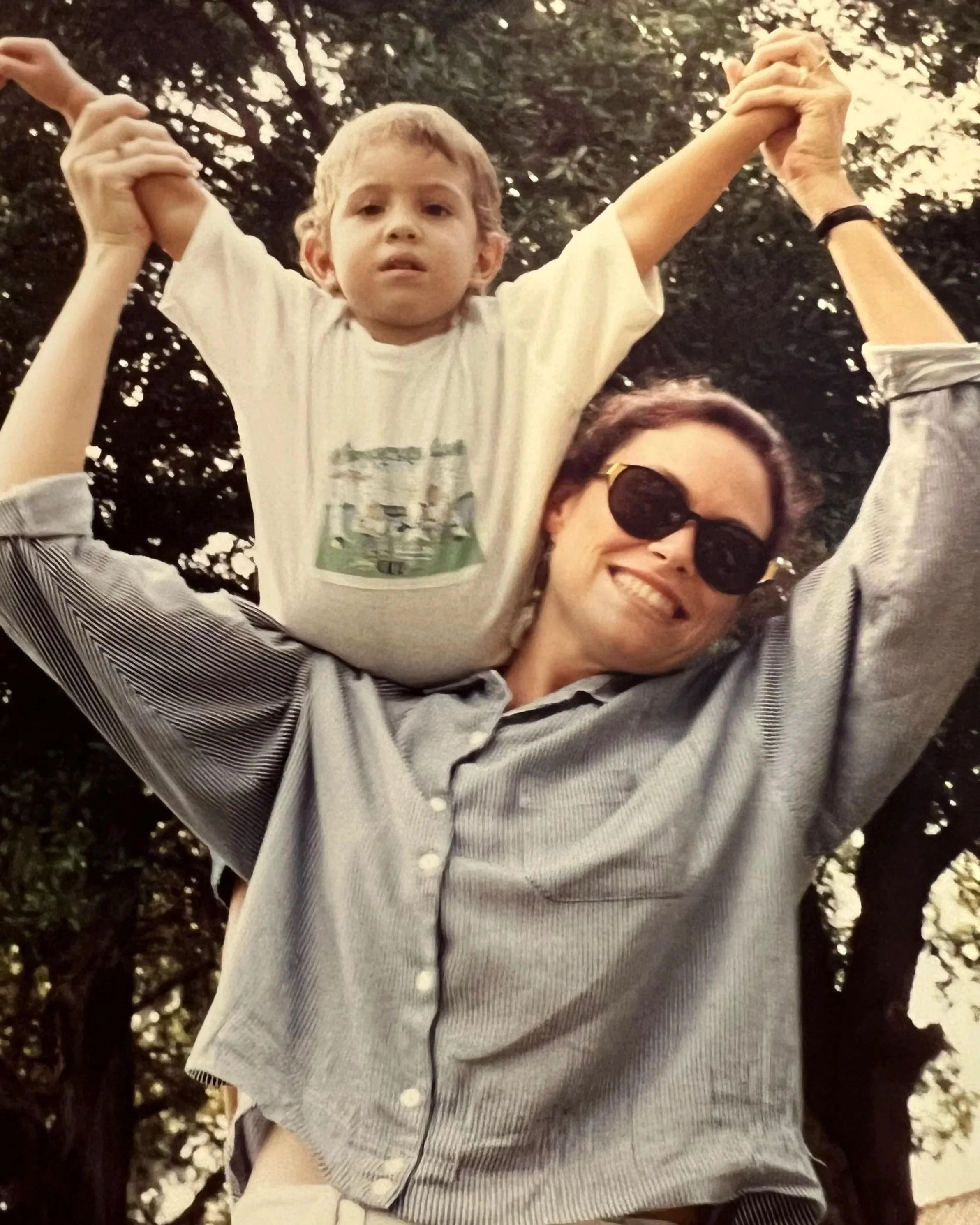 A young boy sitting on an adult woman’s shoulders, holding her hands above his head. The woman is wearing sunglasses and smiling. They are outdoors with trees and foliage in the background.