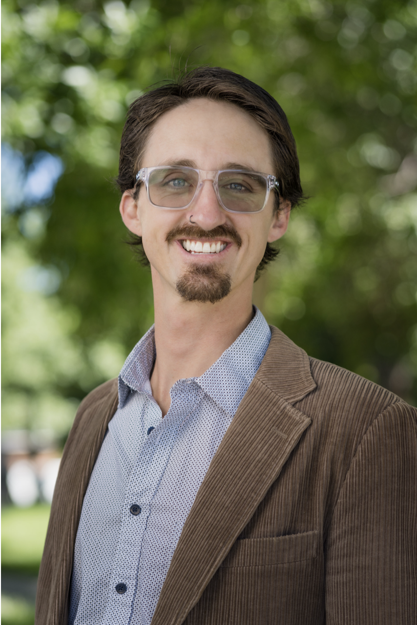 A smiling man with glasses, facial hair, wearing a brown blazer and light blue shirt, standing outdoors with a green, blurred background of trees.