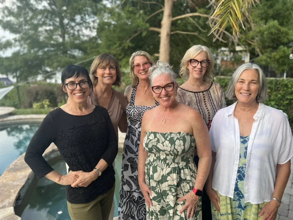 Six middle-aged women posing outdoors by a pool, smiling, with greenery and trees in the background.