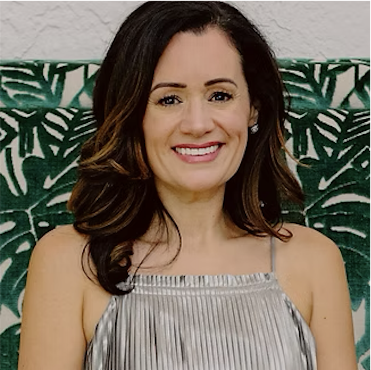 A woman with dark brown hair and a tan dress, smiling in front of green leafy plant pattern.