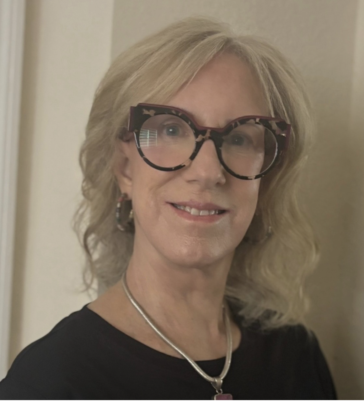 A woman with shoulder-length blonde hair, wearing large tortoiseshell glasses, earrings, a necklace, and a black top, smiling at the camera in front of a beige background.