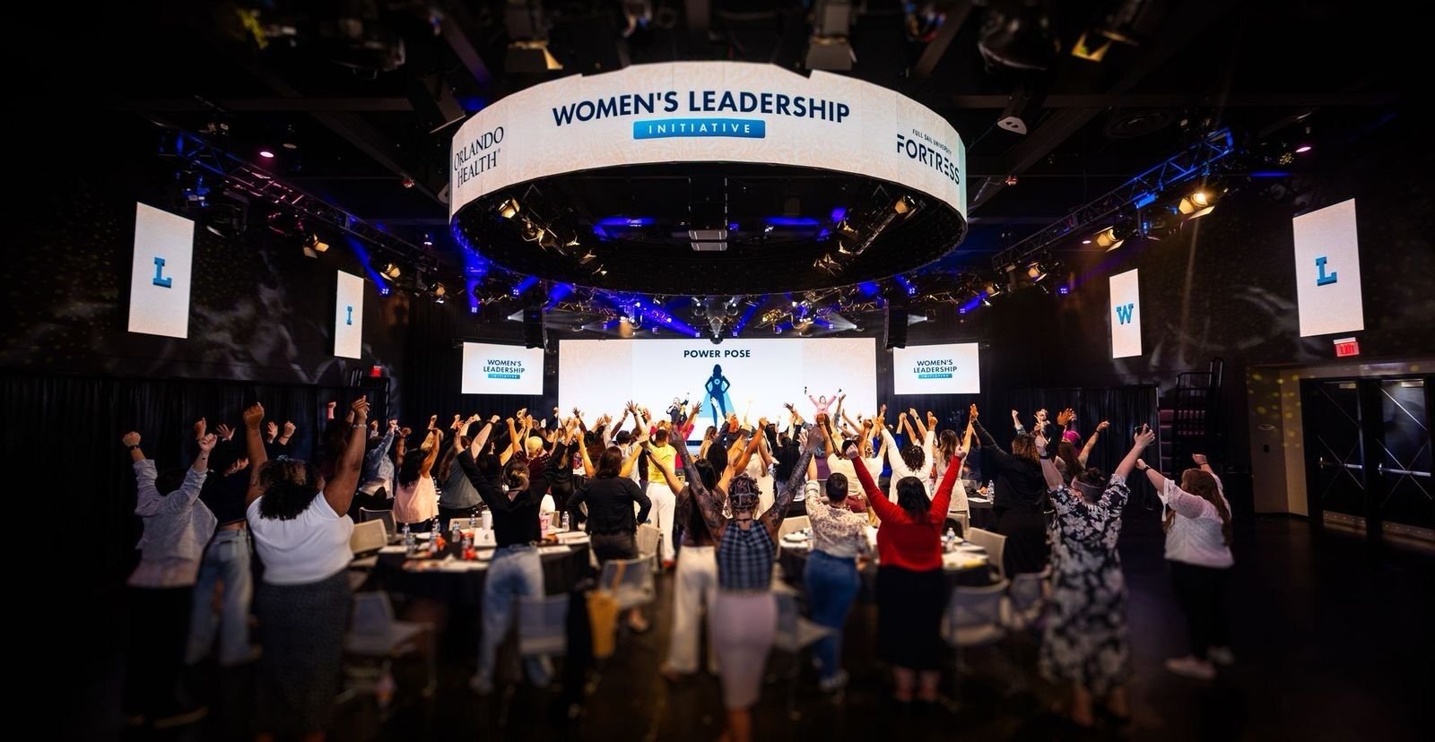A large conference room filled with people standing and raising their hands during a Women's Leadership Initiative event, with a stage displaying a slide titled "Power Pose."