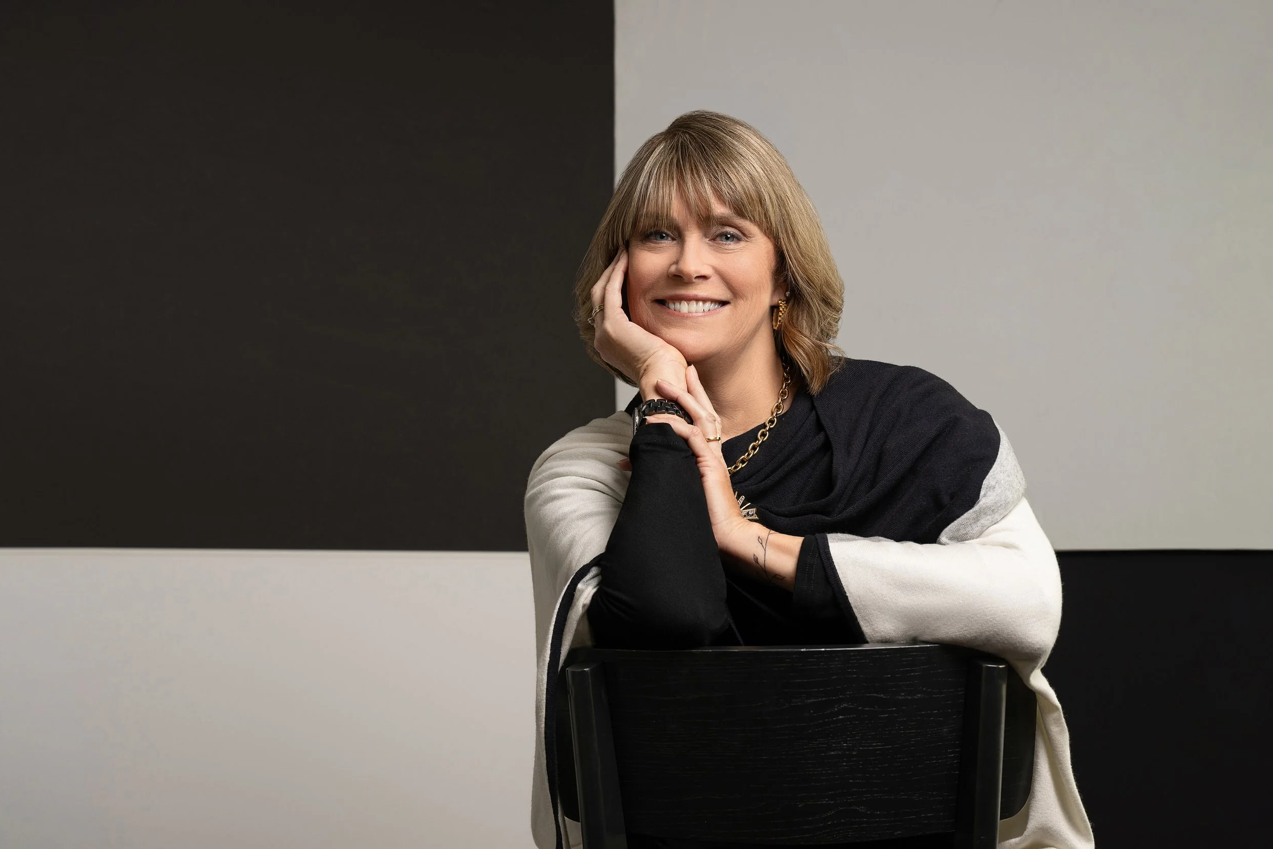 A woman with light brown hair and bangs, smiling and resting her chin on her hand, sitting on a black chair against a background divided into black and gray sections.