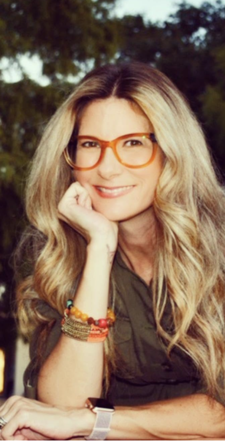 A woman with long, wavy blonde hair wearing orange glasses and a dark top, smiling with her hand resting on her chin, wearing colorful bracelets, outdoors with trees in the background.