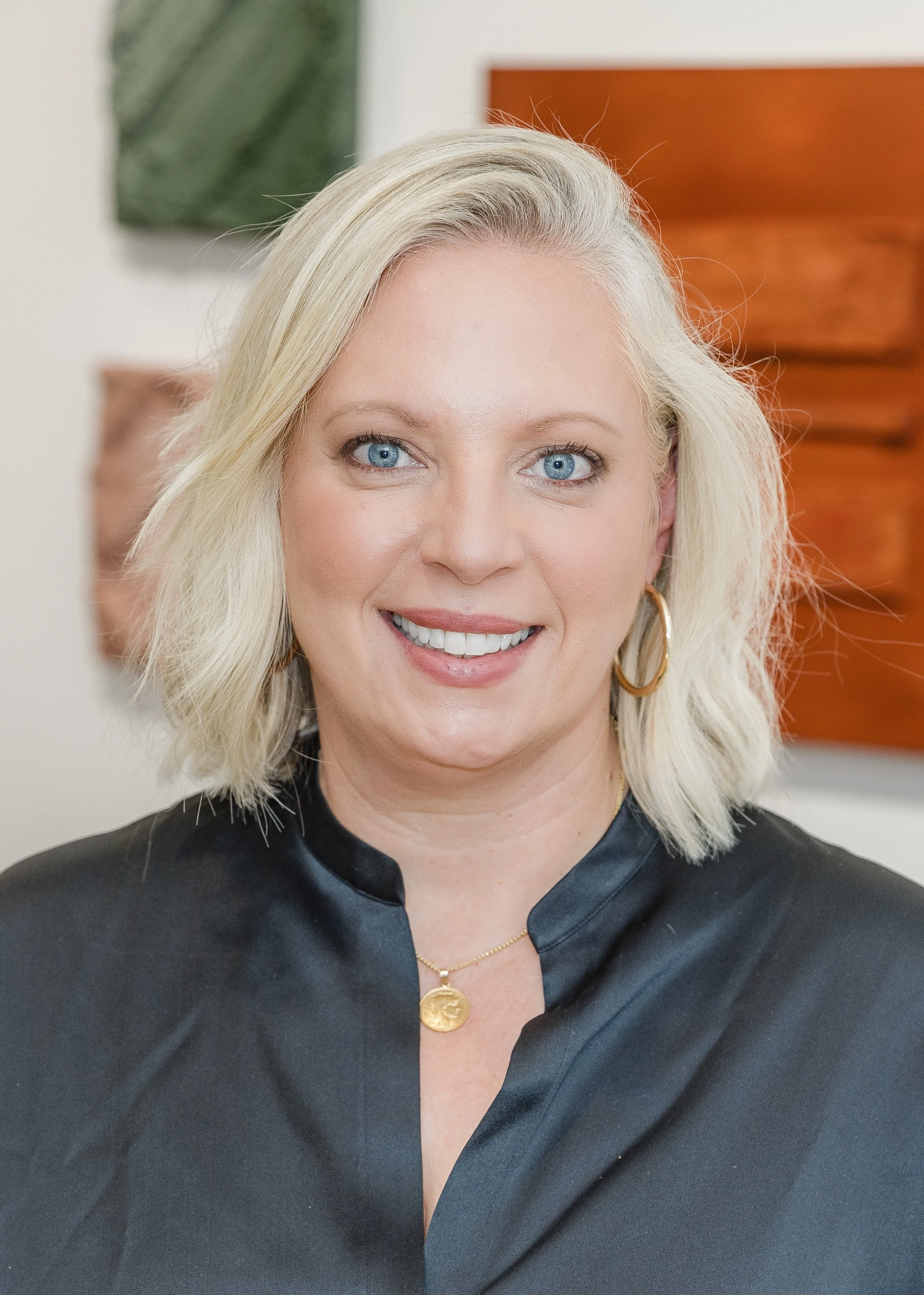 A woman with blonde hair smiling, wearing a black satin-like jacket, gold hoop earrings, and a gold pendant necklace, standing indoors with abstract artwork in the background.