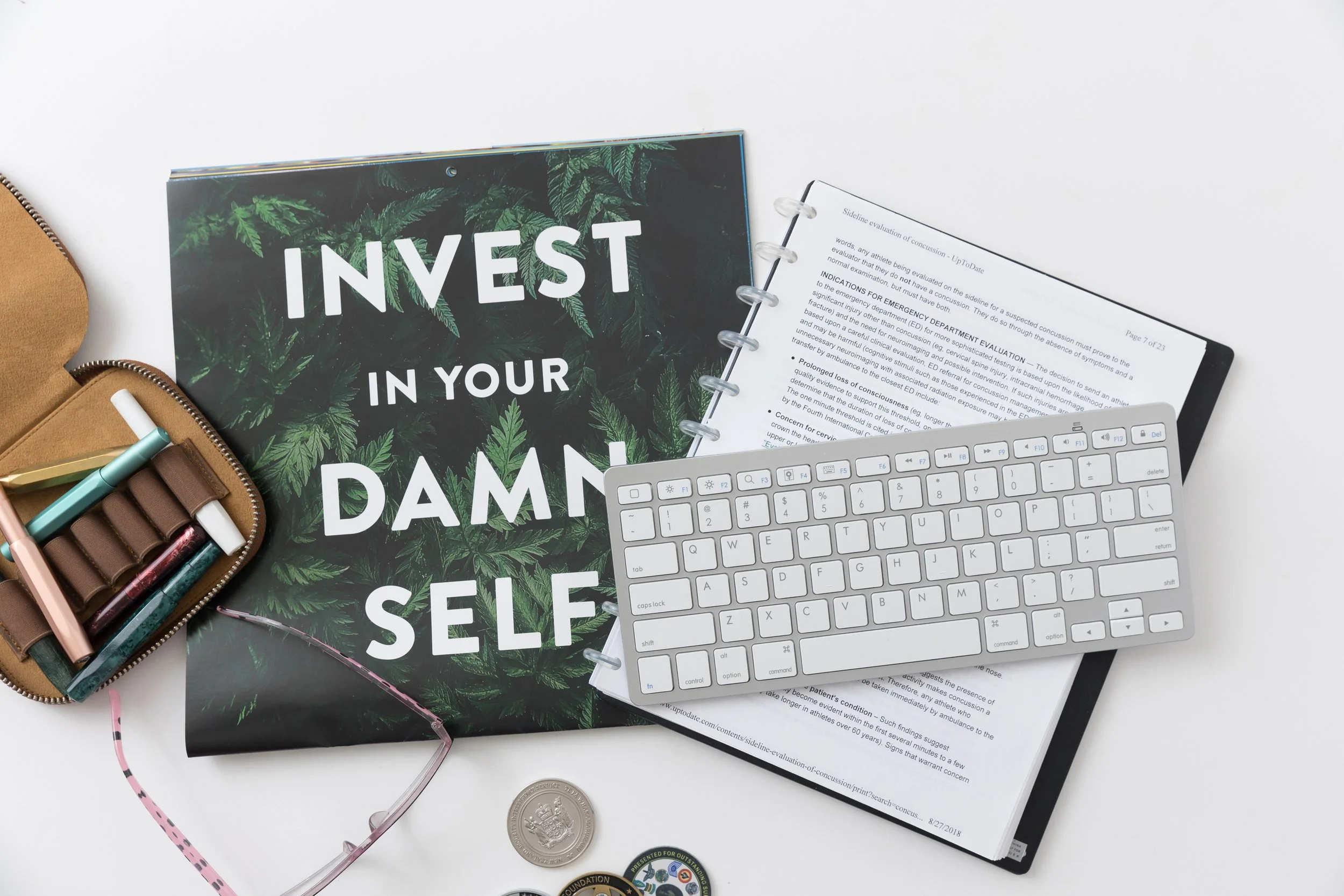 A workspace with a black notebook titled 'Invest in Your Damn Self' with green leafy patterns, an open binder with printed documents, a white computer keyboard, a brown zippered pouch with colored pens, pink and white glasses, a coin, and some round pins on a white surface.
